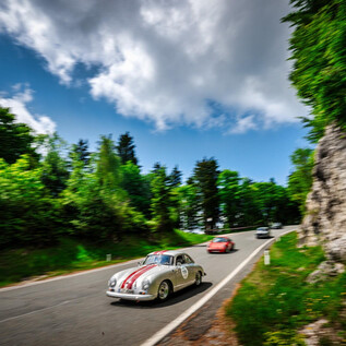 A classic car with racing stripes drives along a winding road. Surrounded by green trees and a blue sky with clouds. | © motorsportpics.at