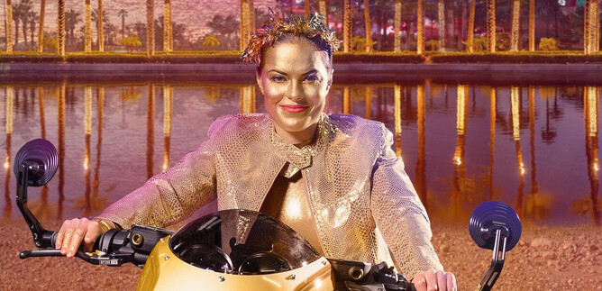 A smiling person sits on a futuristic motorcycle in front of a colorful, illuminated landscape. The background shows trees and reflective water. | © Salzburger Landestheater