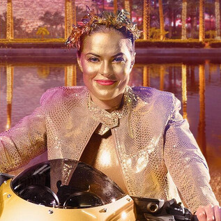 A smiling person sits on a futuristic motorcycle in front of a colorful, illuminated landscape. The background shows trees and reflective water. | © Salzburger Landestheater