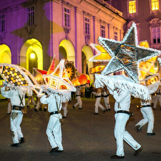 A festive parade with dancers in white costumes carrying large, glowing stars. The backdrop is illuminated by colorful lights. | © Günter Breitegger