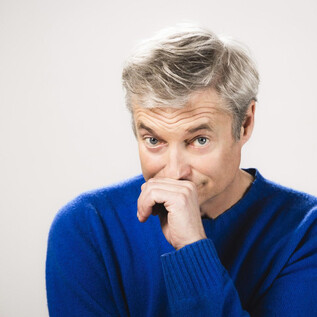 A man with gray hair is wearing a blue sweater. He has a thoughtful expression and is holding his finger to his lips. | © Moritz Schell