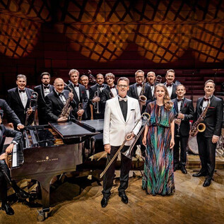 A large jazz band is on a stage, all members are dressed in formal attire. In the foreground, a piano and a singer can be seen. | © Schmidt & Salden