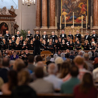 A choir is singing in a large church with many spectators. Behind the choir, a beautiful altar is visible, decorated with candles and artworks. | © Dom zu Salzburg