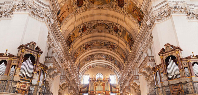 An impressive church interior with high vaults and elaborate ceiling paintings. On both sides, there are large organs, and elegant light illuminates the space. | © Erzdiözese Salzburg