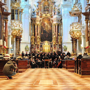 A concert in a magnificent church with richly decorated columns and an impressive altar. The audience sits on wooden benches and listens to the performance. | © P. Alkuin Schachenmayr