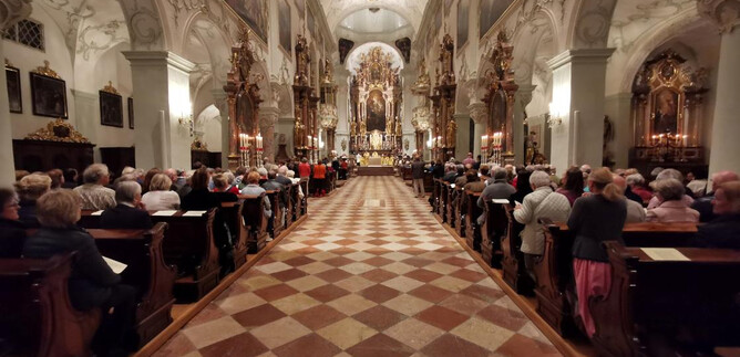A magnificent church with a beautiful ceiling and many decorative details. Many people are sitting in the pews and listening to an event. | © Erzabtei St. Peter
