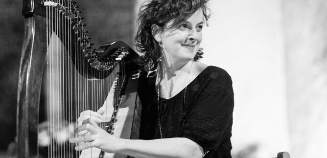A woman is playing the harp and smiling. The background is blurred and contributes to the musical atmosphere. | © Weltenklang