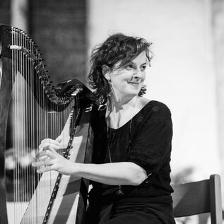 A woman is playing the harp and smiling. The background is blurred and contributes to the musical atmosphere. | © Weltenklang