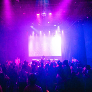 A lively party with many people in the foreground.  
Colorful lights and a DJ stage create an exciting atmosphere. | © Angelina Widmann