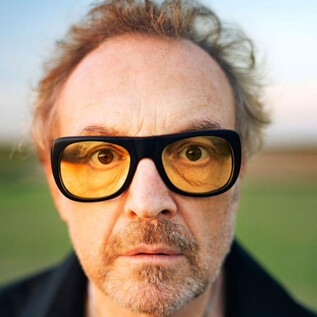 A man with glasses and styled hair looks directly into the camera. In the background, a green landscape can be seen. | © www.lukasbeck.com