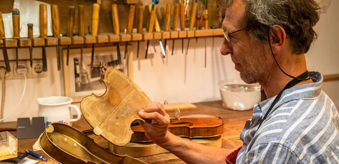 A violin maker inspects the inner sound body of a violin in his workshop. Various tools and additional instruments are visible in the background. | © Andreas Kolarik