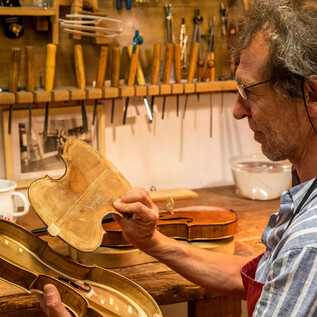 A violin maker inspects the inner sound body of a violin in his workshop. Various tools and additional instruments are visible in the background. | © Andreas Kolarik