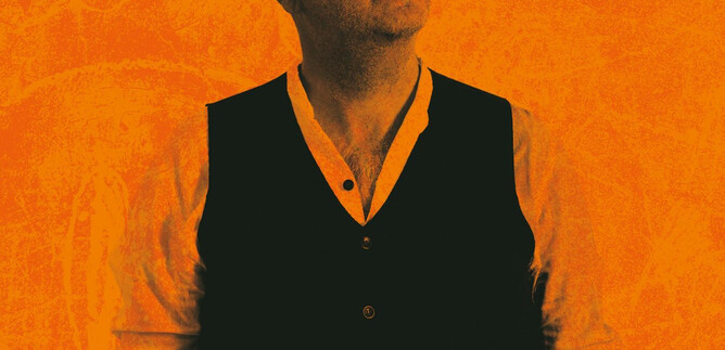 A man with a bald head and sunglasses stands in front of an orange background. He is wearing a black vest and looking up. | © Tobias Koark-Haberl