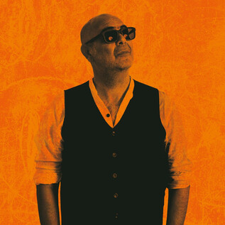 A man with a bald head and sunglasses stands in front of an orange background. He is wearing a black vest and looking up. | © Tobias Koark-Haberl