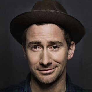 A man with a hat smiles at the camera. The background is dark and plain. | © Christian Brecheis