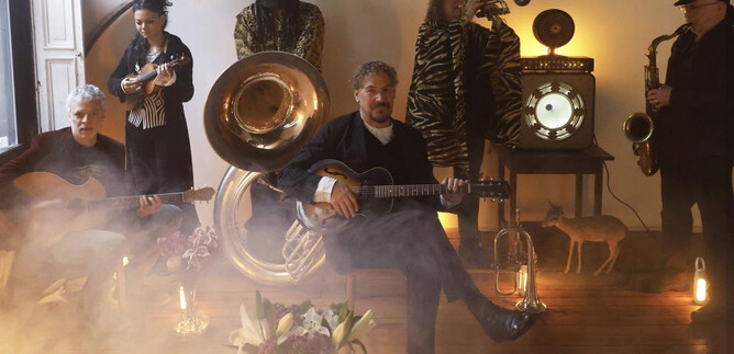 A band plays in a room while a mystical atmosphere is created with fog and soft lights. In the foreground, a musician sits with a guitar and the other instrumentalists stand in the background. | © Agentur/Band