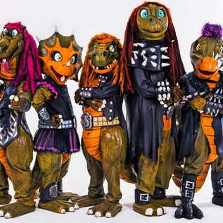 A group of dinosaur mascots in rockin' outfits stands in front of a dramatic background landscape. The text "Heavy Saurus" is prominently placed. | © sonymusic.de