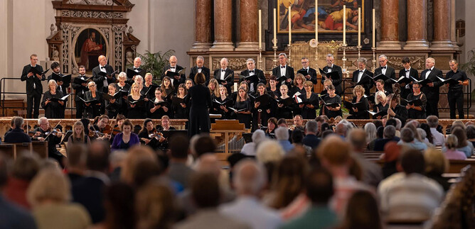 A choir is standing on a stage in a church and singing. In the background, the altar and numerous spectators can be seen. | © Dom zu Salzburg