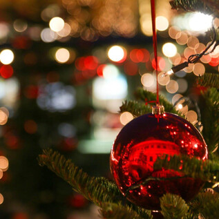 A red Christmas tree ornament hangs on a tree. Bright lights and Christmas decorations shine in the background. | © Hellbrunner Adventzauber