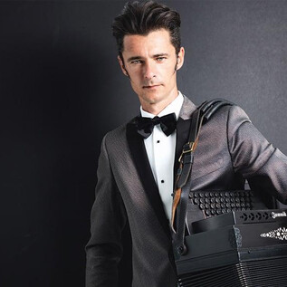 A well-dressed man is holding a black accordion and standing in front of a neutral background. He is wearing a tuxedo with a bow tie and has a confident expression. | © Günther Egger