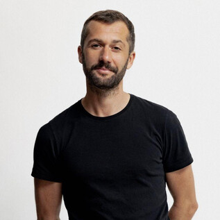 A man stands in front of a neutral background. He is wearing a black t-shirt and has a light beard. | © Susan Buth