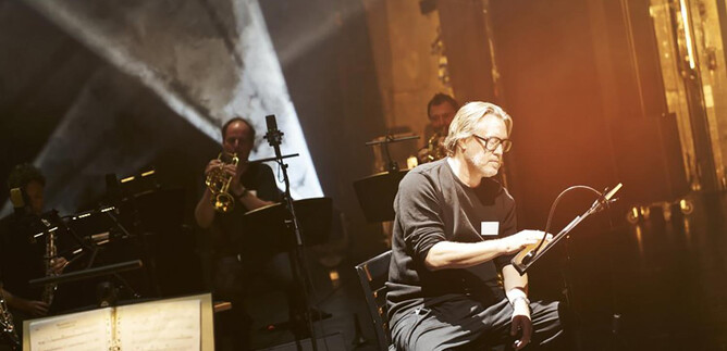 A musician is sitting on stage and working on a sheet of music. In the background, other musicians with instruments are visible. | © Tommy Hetzel