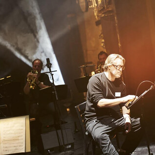 A musician is sitting on stage and working on a sheet of music. In the background, other musicians with instruments are visible. | © Tommy Hetzel