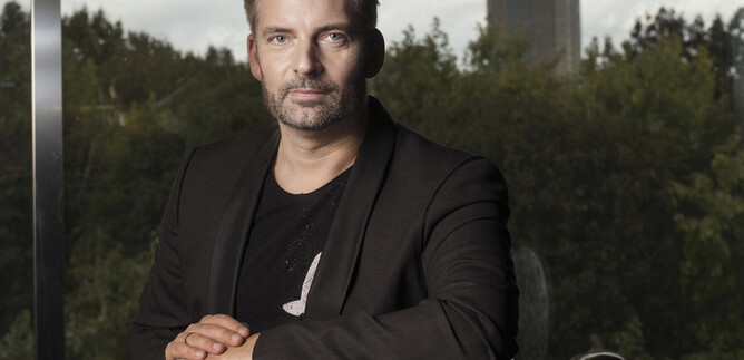 A man in a black blazer is sitting at a table and looking directly at the camera. In the background, trees and a high-rise building are visible. | © Franck Ferville