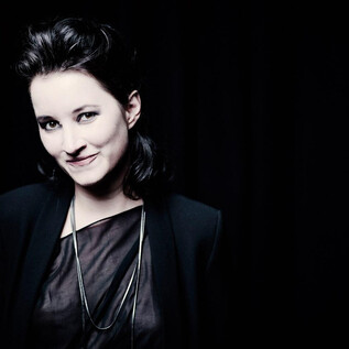 A woman with dark hair smiles at the camera. The background is black, which draws the focus to her face. | © Marco Borggreve