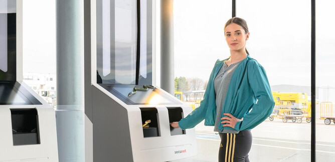 A woman stands at modern self-service terminals at the airport. In the background, there are windows overlooking the airport operations. | © SLT / Christian Krautzberger