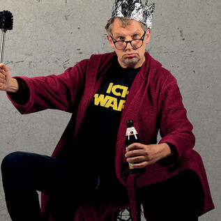 A man in a red bathrobe is wearing an aluminum hat and holding a bottle of beer in one hand and a broom in the other. He is sitting with a confident expression in front of a gray background. | © Dominic Reichenbach