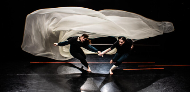 Two dancers move dynamically across a dark surface, while a light fabric sways above them. The image radiates energy and movement. | © Fabian Schober