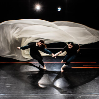 Dvě tanečnice dynamicky tančí na tmavém povrchu, zatímco nad nimi vlaje lehká tkanina. Obraz vyzařuje energii a pohyb. | © Fabian Schober