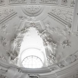 A choir stands in a circular arrangement in a historic room. The singers are wearing black clothing and are focused on the conductor. | © Stefan Zenzmaier & Kollegienkirche
