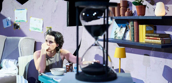 A person sits pensively at a table in a simply furnished room. Behind them are bookshelves, and an hourglass is visible in the foreground. | © Tom Leather Neumeier
