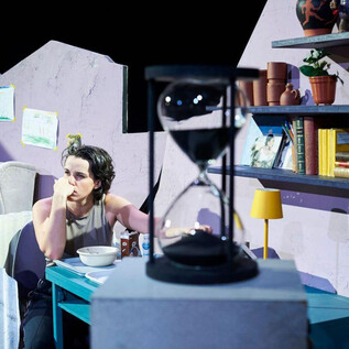 A person sits pensively at a table in a simply furnished room. Behind them are bookshelves, and an hourglass is visible in the foreground. | © Tom Leather Neumeier