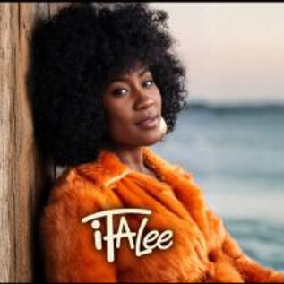 Una mujer con cabello rizado lleva un abrigo naranja y se apoya en una pared de madera. En el fondo se puede ver el mar y el cielo. | © Jazzit