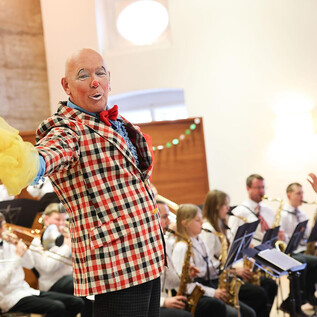 A cheerful clown in a checkered suit stands in the foreground entertaining the audience. In the background, an orchestra plays with various instruments. | © Franz Neumayr