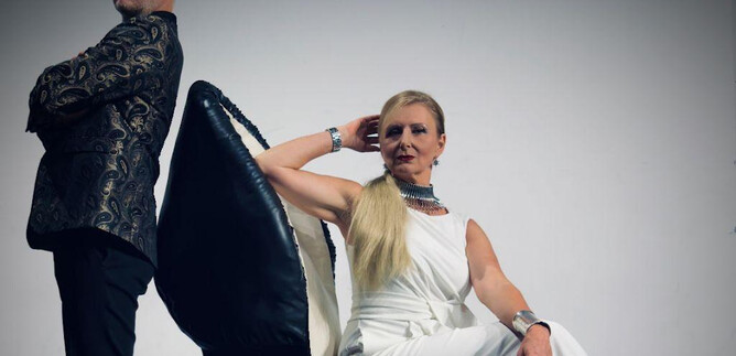 A man and a woman are posing in front of a simple background. The woman is sitting on an oversized shoe and is wearing an elegant white outfit. | © Beatriz Teixeira