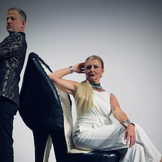 A man and a woman are posing in front of a simple background. The woman is sitting on an oversized shoe and is wearing an elegant white outfit. | © Beatriz Teixeira