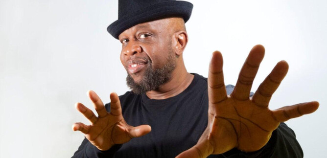 A man in a black shirt and a hat is making a striking gesture with his hands. He has a friendly smile on his face. | © Jazzit