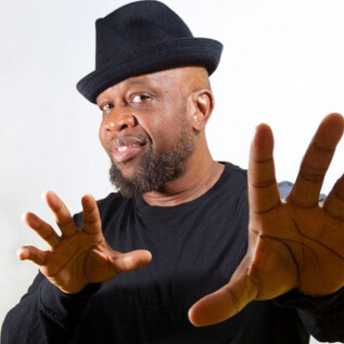 A man in a black shirt and a hat is making a striking gesture with his hands. He has a friendly smile on his face. | © Jazzit