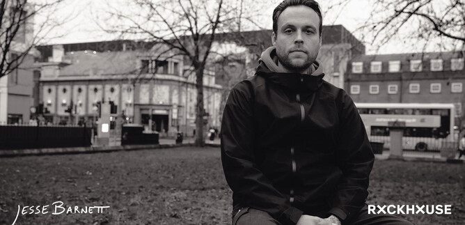 A man is sitting on a meadow in an urban environment. In the background, buildings and trees are visible, conveying a grey atmosphere. | © Rockhouse Salzburg