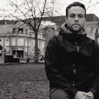 A man is sitting on a meadow in an urban environment. In the background, buildings and trees are visible, conveying a grey atmosphere. | © Rockhouse Salzburg