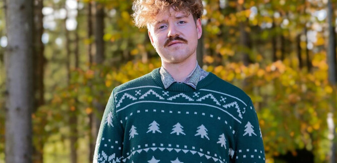 A young man with curly hair is wearing a green Christmas sweater. He stands in the woods in front of colorful autumn trees. | © Anna Sophie Koelbl