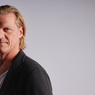 A man with longer blonde hair and a black blazer stands in front of a neutral background. He looks directly at the camera and appears confident. | © Jeff Mangione