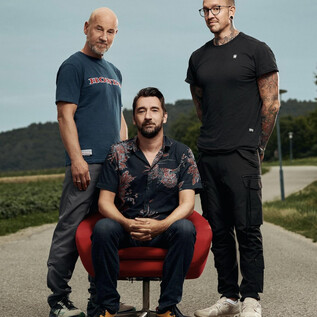Three men are standing on a street. The man in the middle is sitting on a red chair, while the other two are standing and posing in fashionable clothes. | © Pascal Riesinger