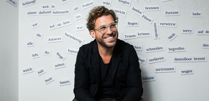 A smiling man sits in front of a wall covered with many written words. He is wearing a black jacket and glasses. | © Chris Gonz