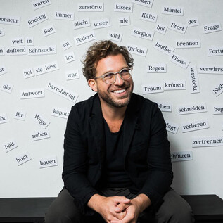 A smiling man sits in front of a wall covered with many written words. He is wearing a black jacket and glasses. | © Chris Gonz