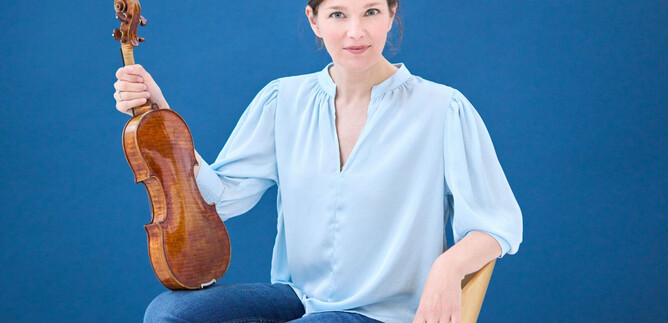 A woman is sitting on a chair and holding a violin in her hand. The background is blue and the woman is wearing a light blue top. | © Kaupo Kikkas